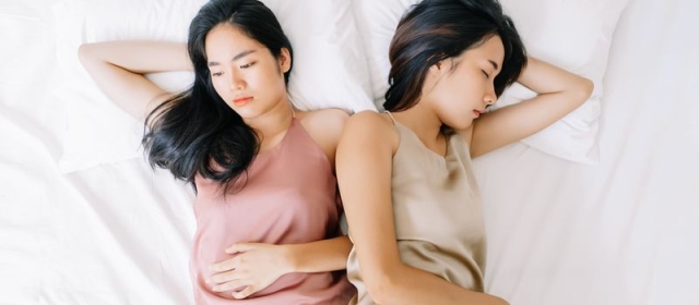 Two women of Asian descent lay next to each other on white sheets. Both wear tank tops. One wears pink, the other wears tan. Both women are looking in opposite directions. 