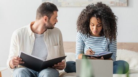 A couple is pictured. The man has short hair, and is wearing a jacket. He sits next to a woman and he is reading a book. The woman is not looking at the man, she is writing in her book instead. the woman has curly hair and wears a blue striped shirt.