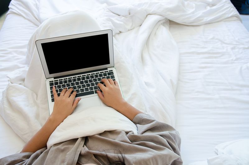 A person, only visible from the chest down, is viewed in their bed. They are covered in a white blanket, sitting on white sheets. They are using a laptop.